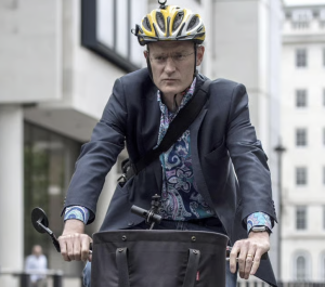 Jeremy Vine wearing blue sportscoat and yellow, white and black bike helmet riding a bicycle.